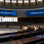 Sala de sessões vazia na CLDF com documentos desorganizados e relógios atrasados, simbolizando lentidão e ineficiência.