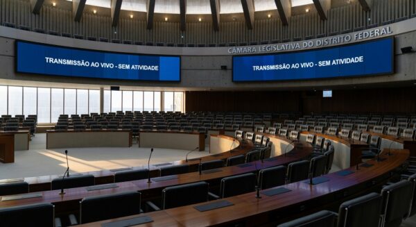 Sala de sessões vazia na CLDF com documentos desorganizados e relógios atrasados, simbolizando lentidão e ineficiência.