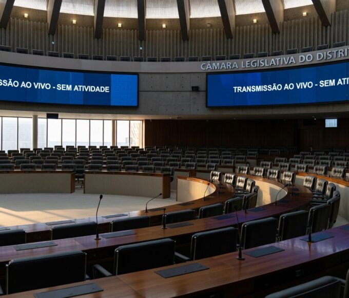 Sala de sessões vazia na CLDF com documentos desorganizados e relógios atrasados, simbolizando lentidão e ineficiência.