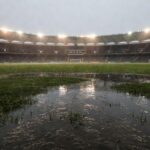 Gramado de futebol alagado por chuva intensa no Candangão, com poças d'água e grama molhada em estádio no DF.
