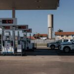 Posto de gasolina no Brasil com preços de combustíveis elevados e viaturas da Polícia Federal ao fundo.